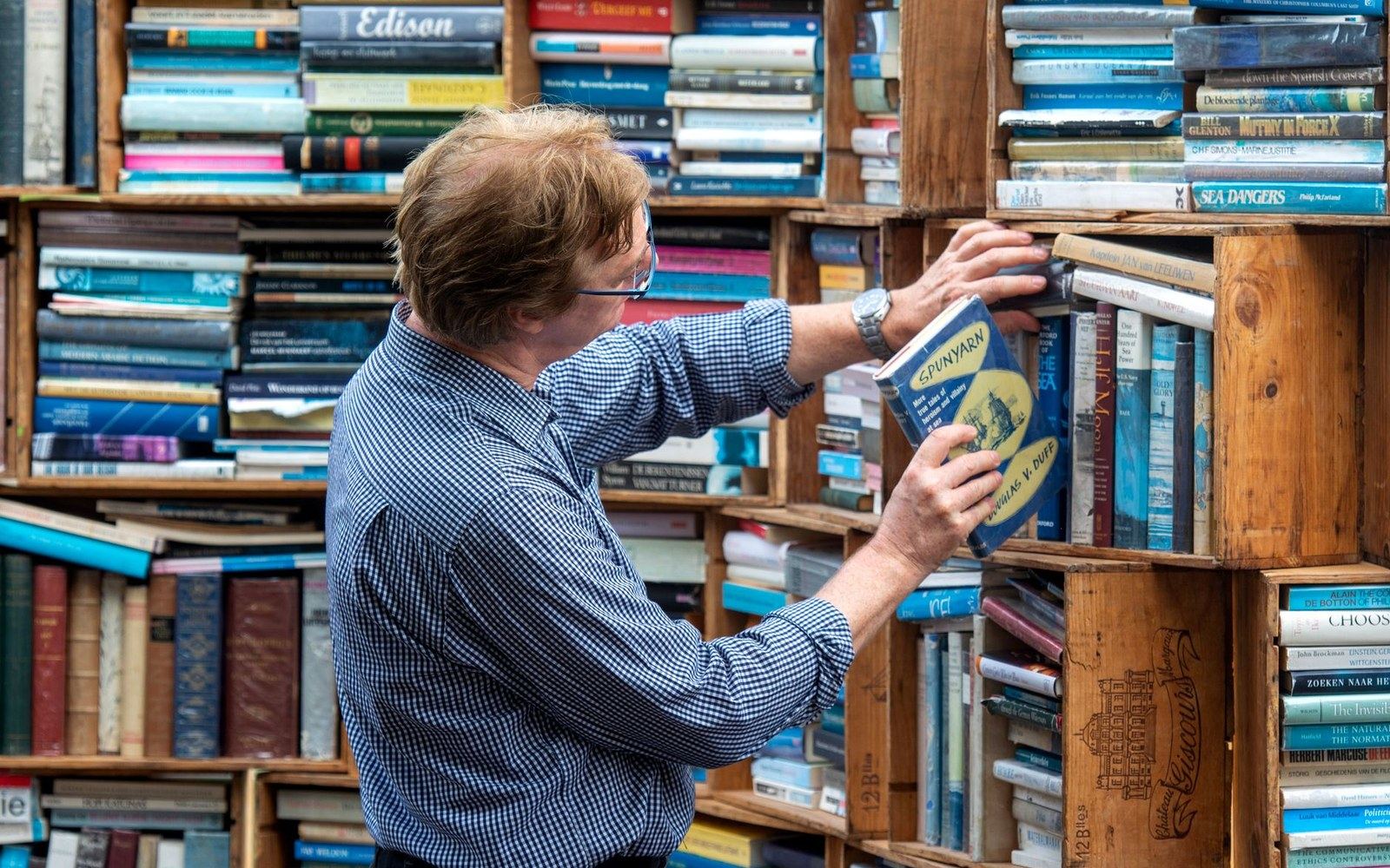 A bookshop display table with Russian-literature translations face-out, an adult browser's hand lifting one title, other spines visible behind
