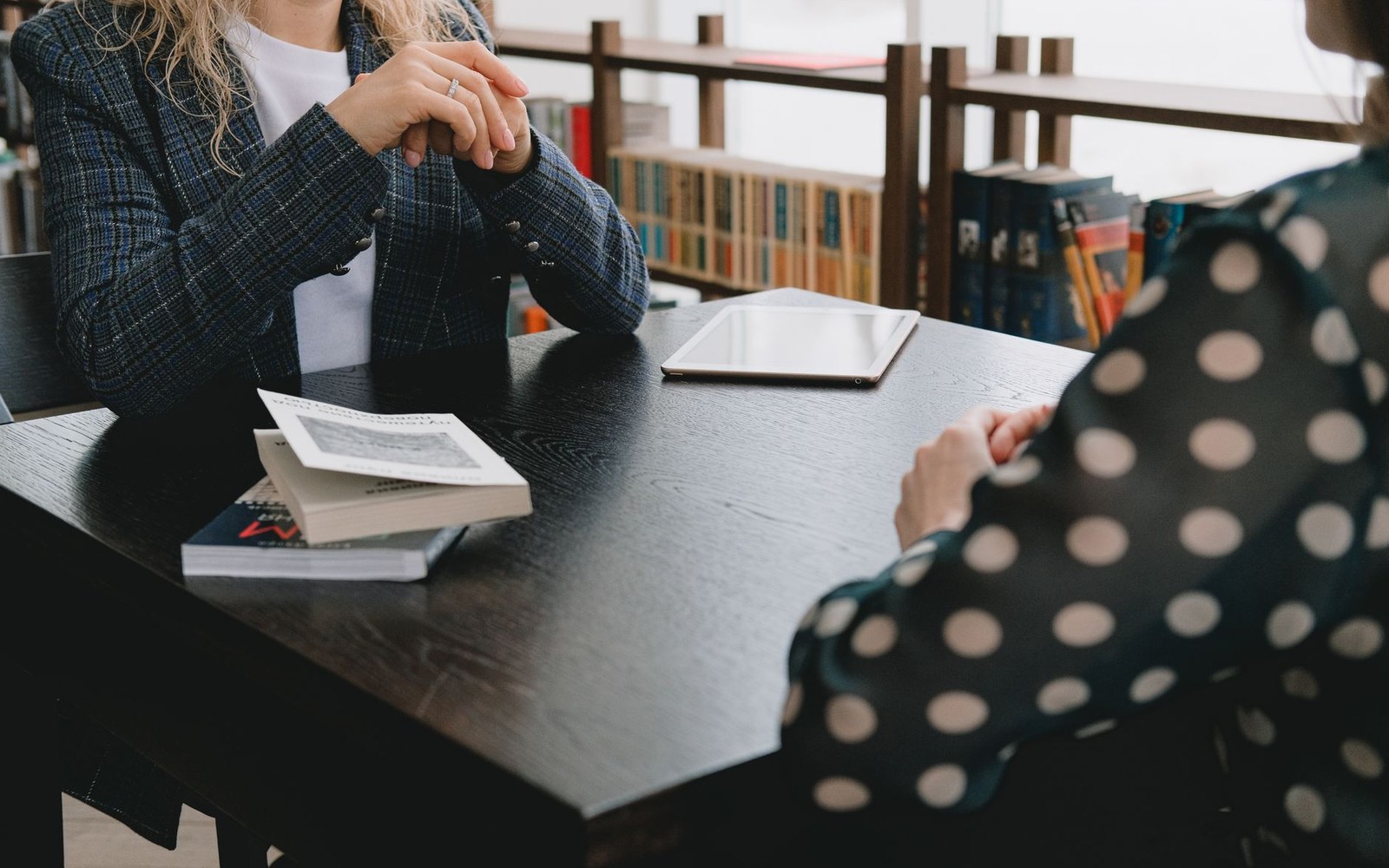 Two women sit across a dark table with books and a tablet during a discussion