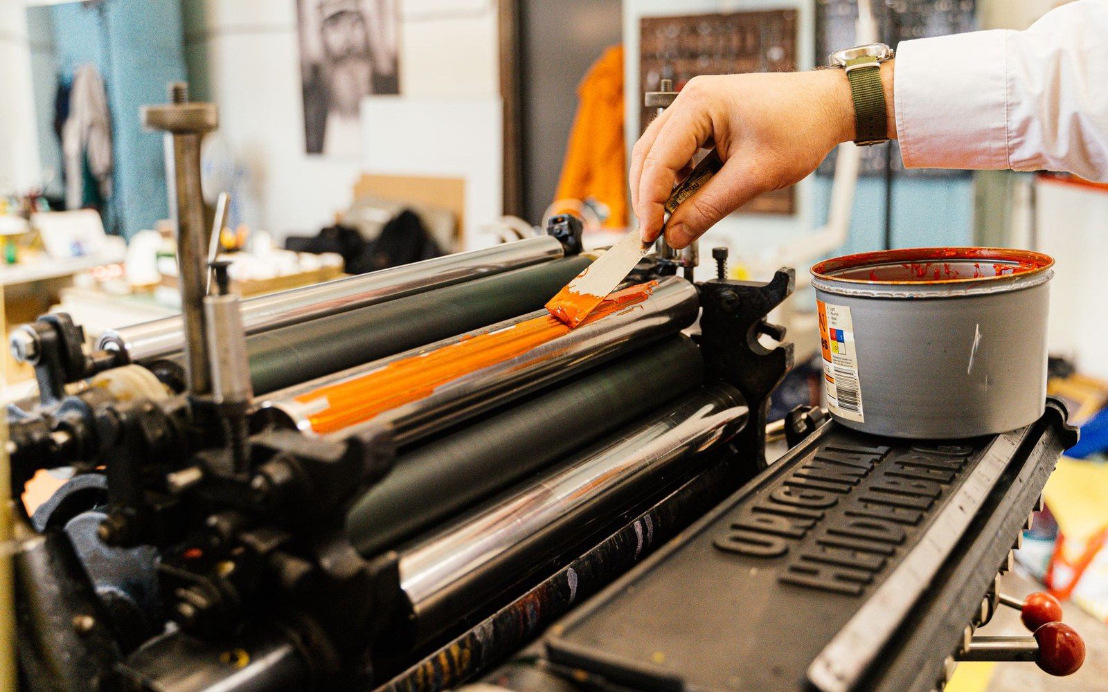 A printing press mid-run, paper moving through, an adult technician adjusting the feed, ink-stained hands in close frame